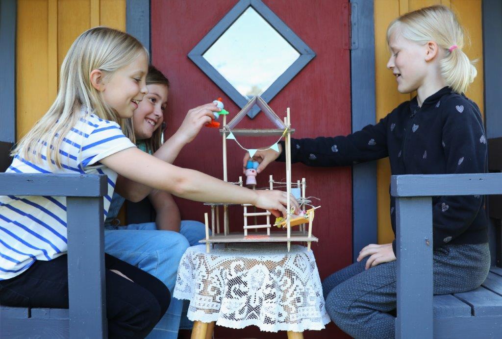 Kolme lasta istuu penkeillä vastakkain. He leikkivät välissä olevalla pöydällä olevalla avoimella nukkekodilla.